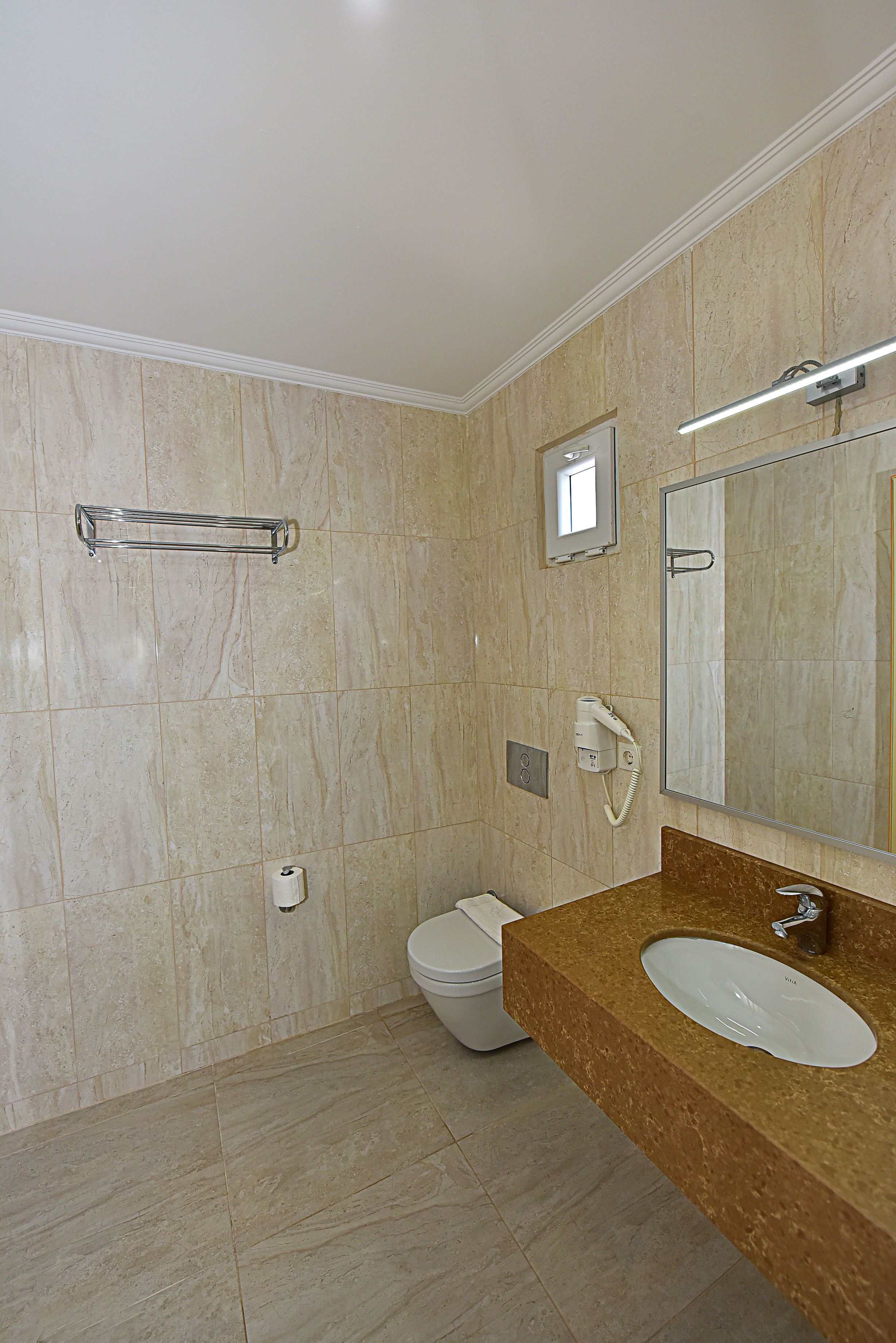 A bathroom with a marble floor, a white toilet, a sink with a mirror, and a hairdryer on the wall.