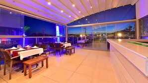 Un comedor interior con mesas y sillas, ventanas amplias y vistas de la ciudad de noche.