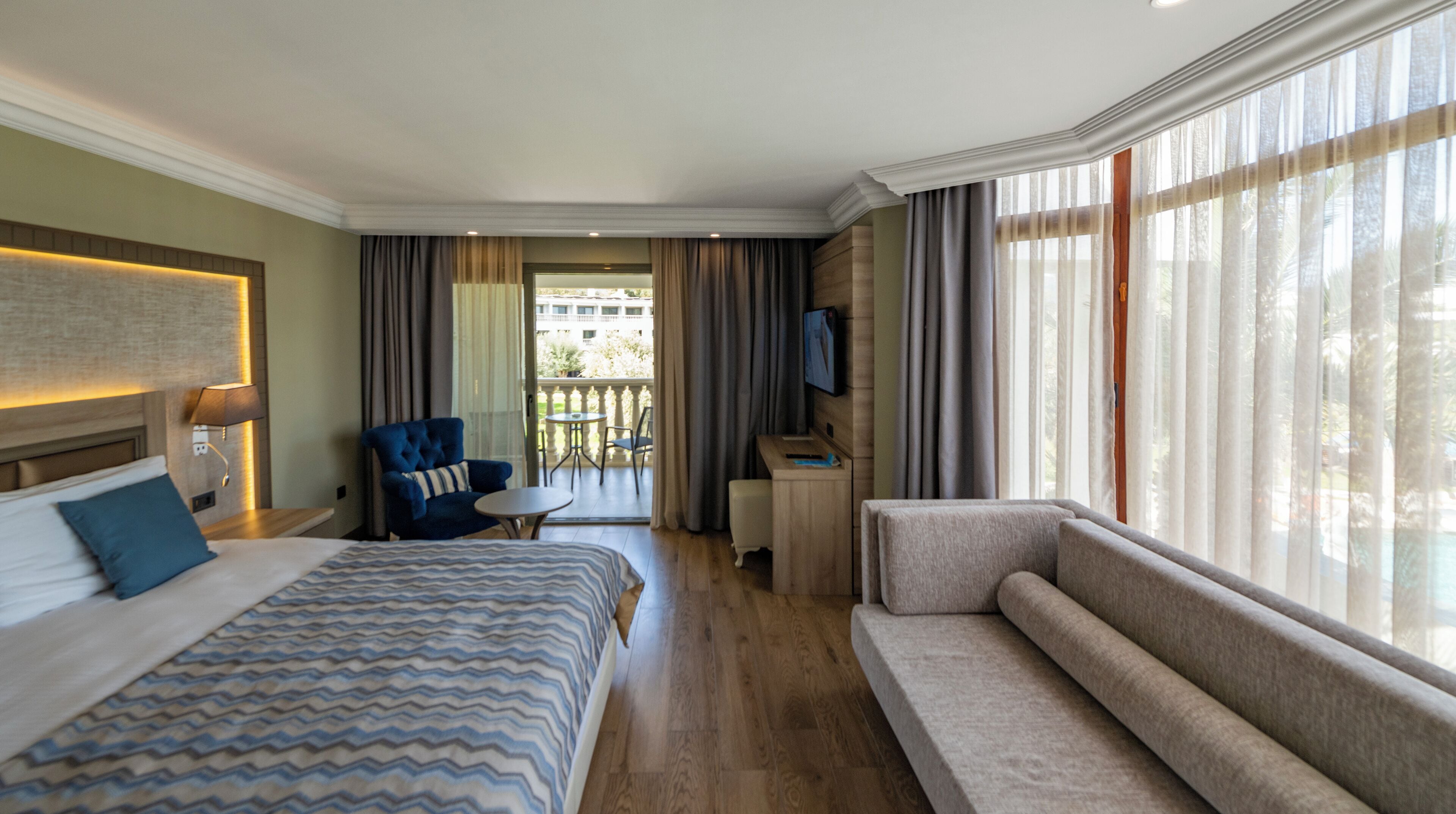Une chambre d’hôtel avec un lit, un canapé, une télévision, un bureau et un balcon avec des rideaux.
