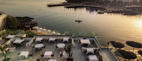 An outdoor dining area with tables and chairs, overlooking a calm body of water with boats and a pier.