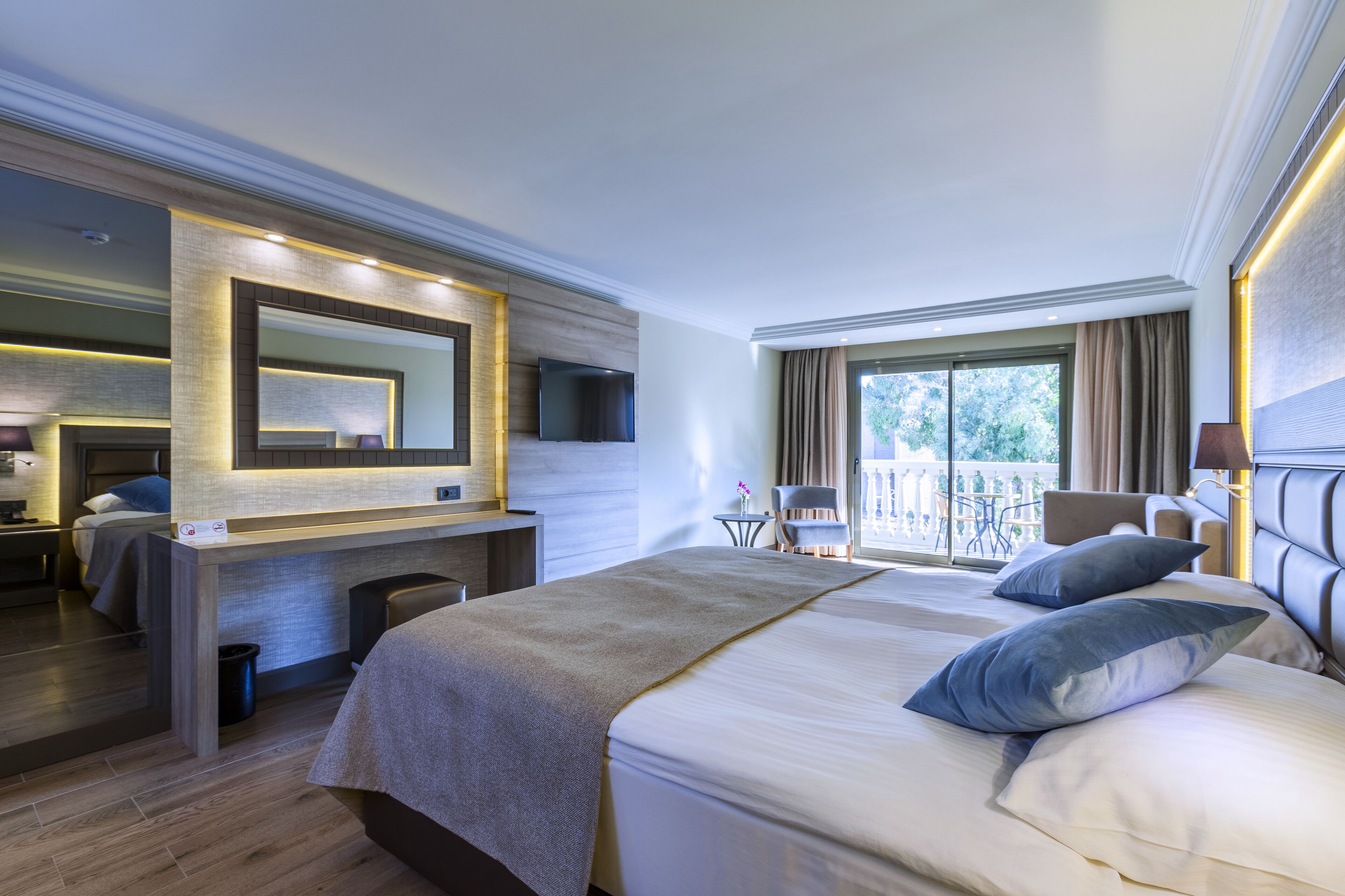 A modern hotel room with a large bed, a desk, a chair, a TV, and a balcony with a view.