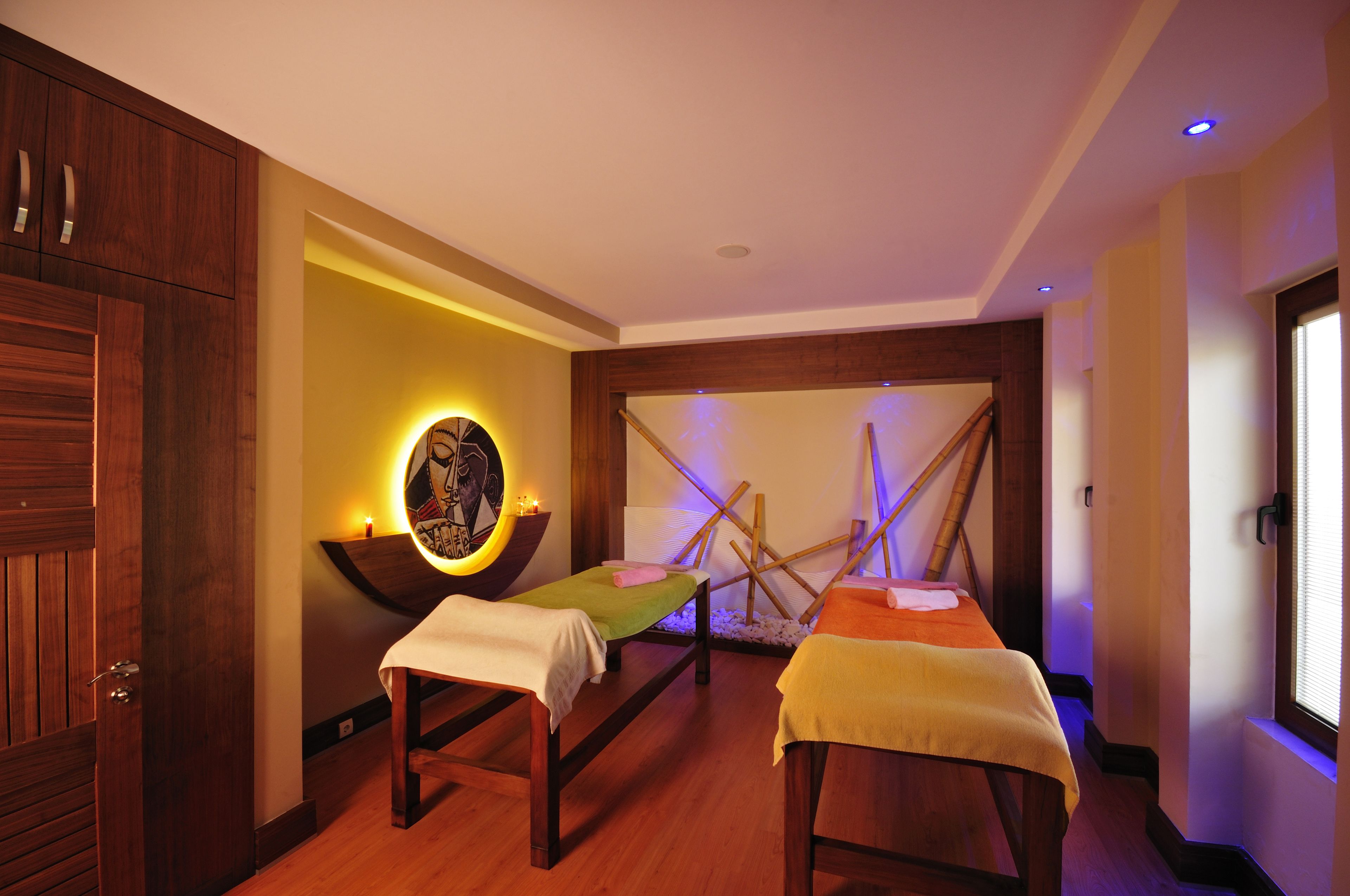 A spa room with two wooden beds, a wall-mounted massage table, and a decorative wall with a circular light fixture.