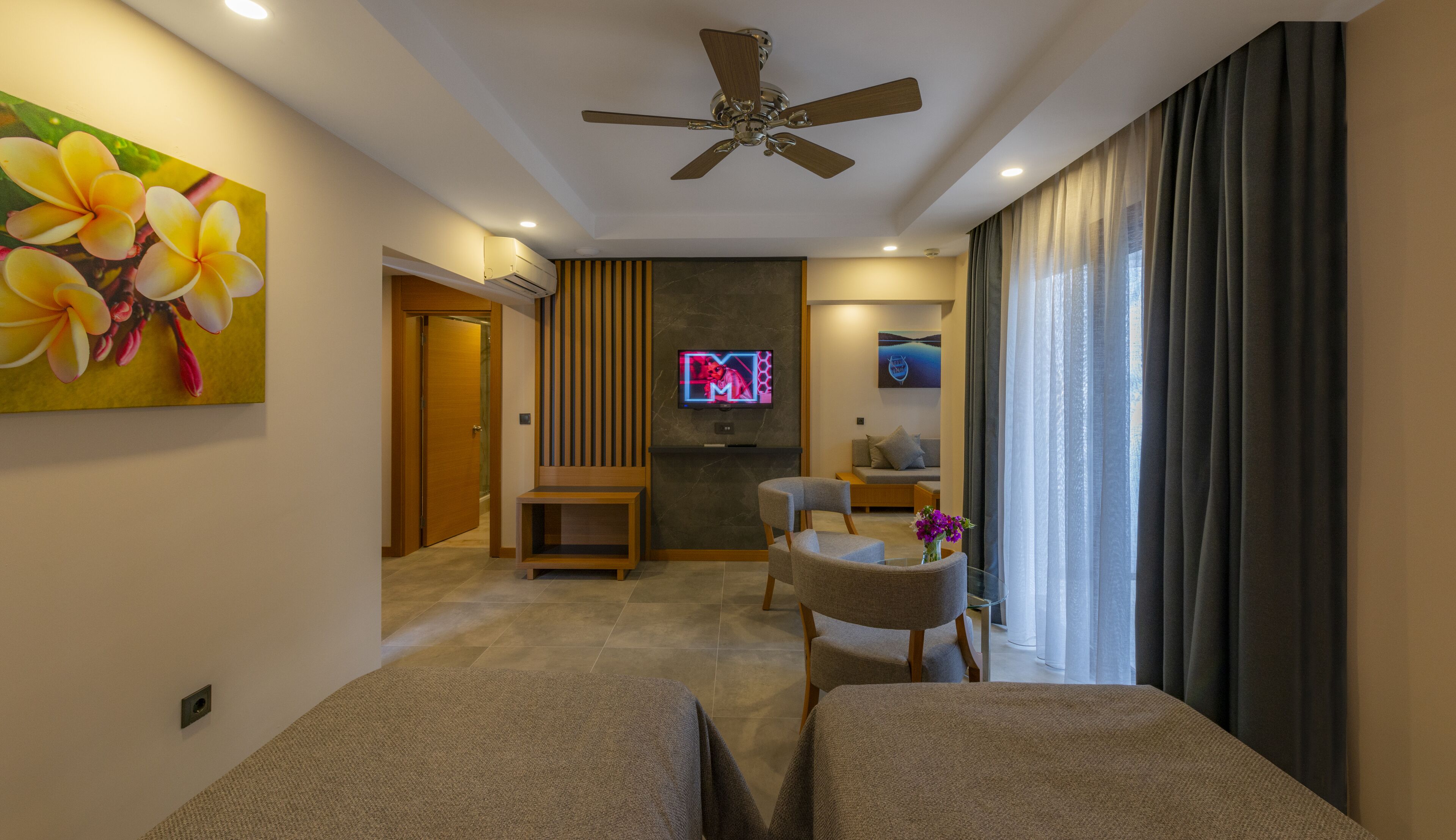 A modern living room with a ceiling fan, a flat-screen TV mounted on a dark wall, a sofa, a coffee table, and a large window with curtains.