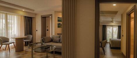A modern hotel room with a sofa, armchairs, a glass coffee table, and a view of the outdoors.
