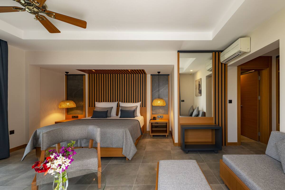 A modern hotel room with a bed, a sofa, a small table with flowers, a ceiling fan, and a mirror.