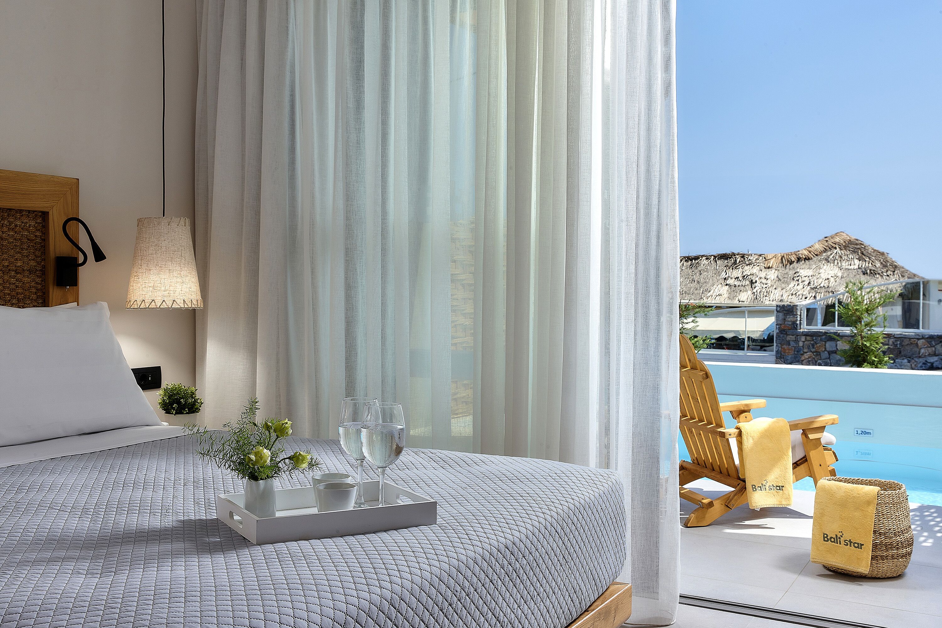 Ein Hotelzimmer mit Bett, Tablett mit Gläsern und Blumen, Stuhl am Pool und Blick auf Pool und Berge.