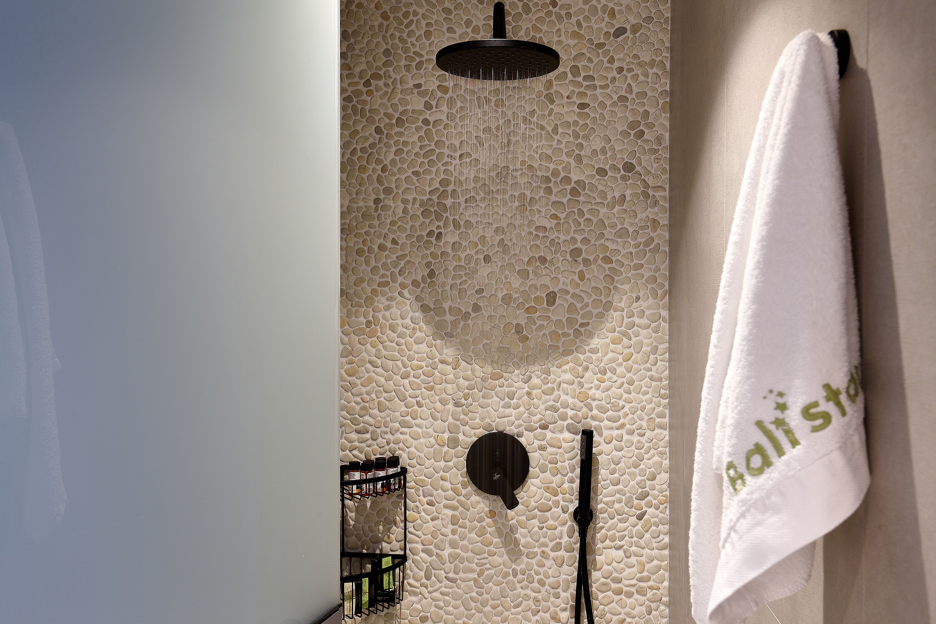 A bathroom with a stone wall, a showerhead, a towel rack with a towel, and a soap dispenser.