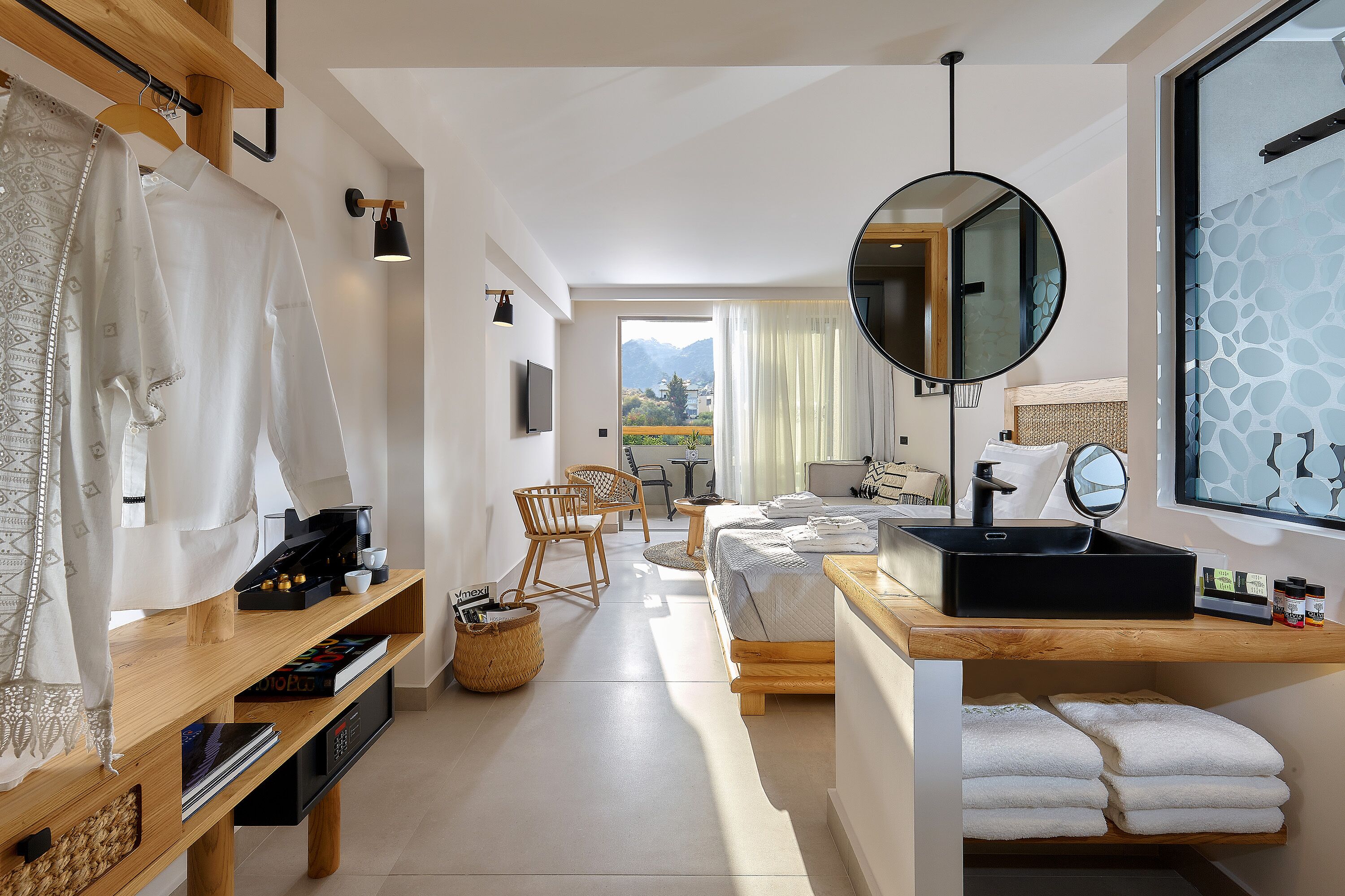 Una camera d'albergo moderna con un letto di legno, un comodino con lavabo nero, uno specchio grande e vista sulle montagne.