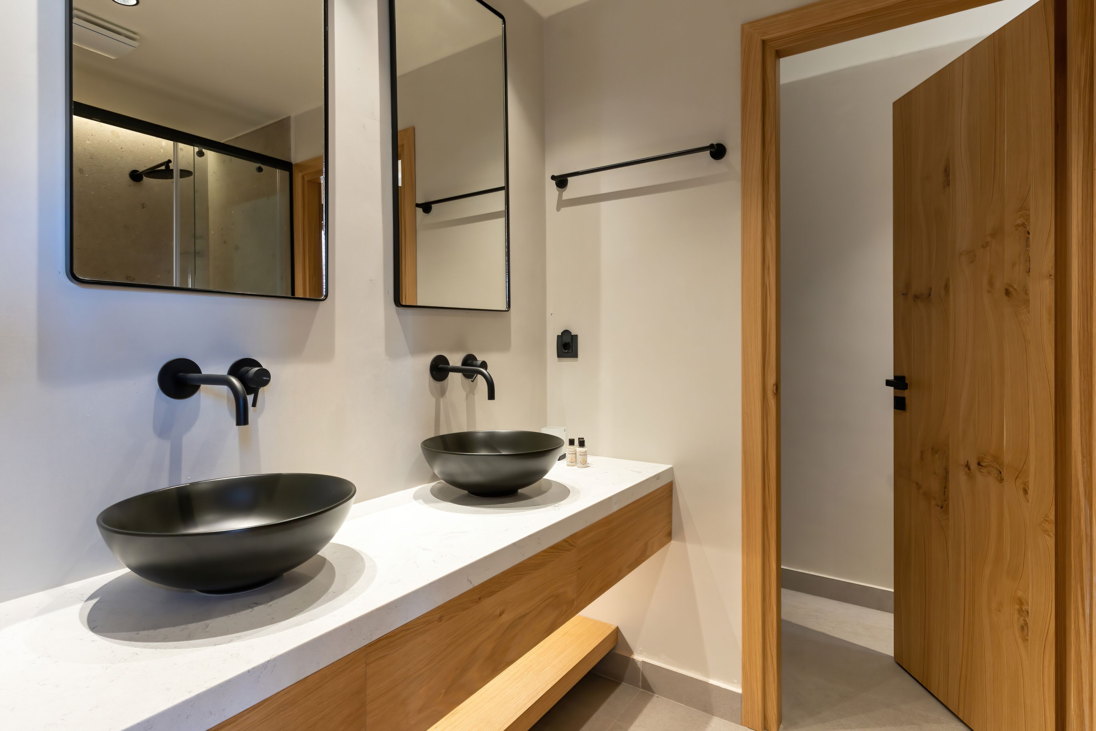 A modern bathroom with a large mirror, two black bowls on a white countertop, and a wooden door.