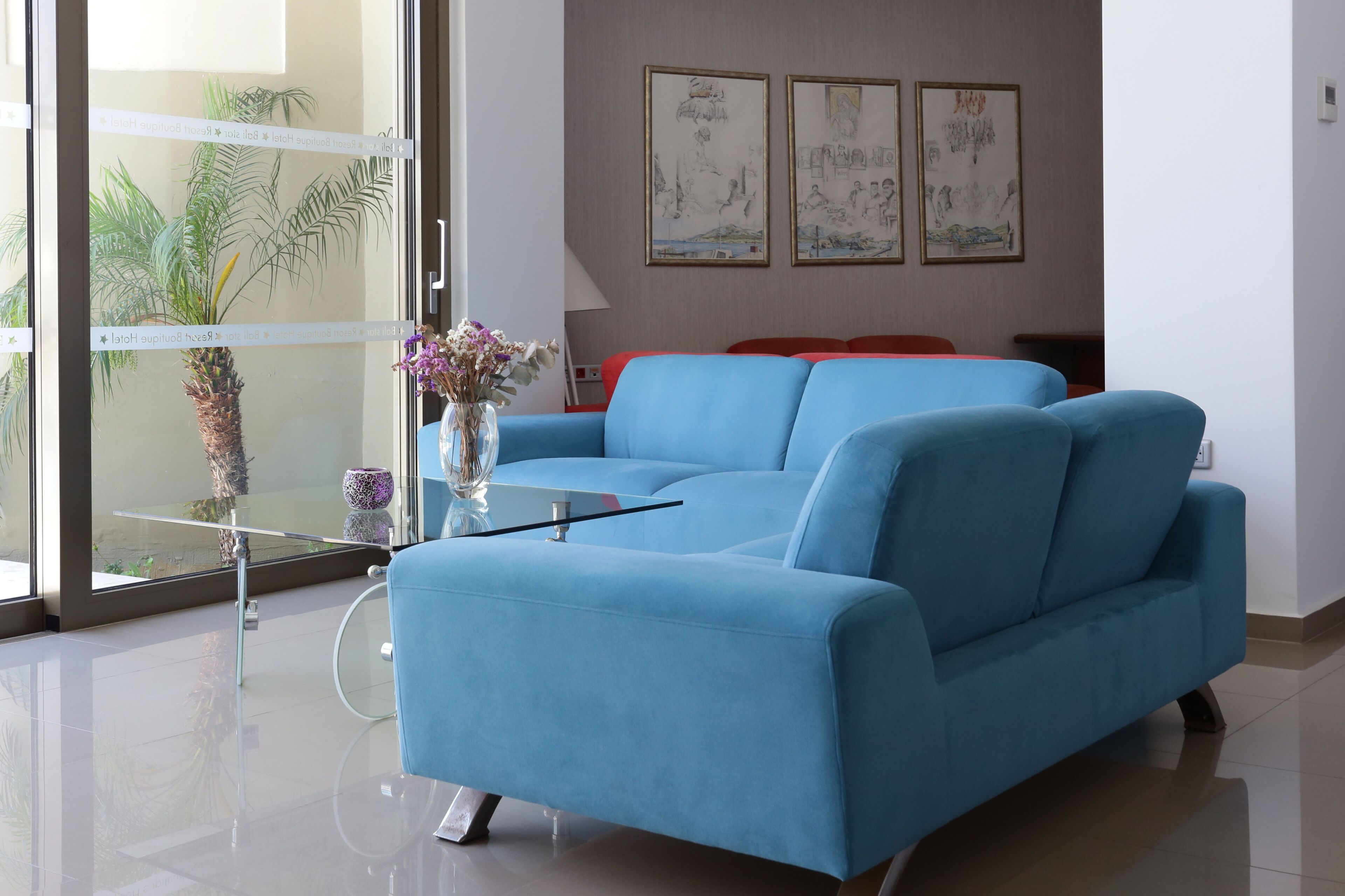Una sala de estar moderna con un sofá azul, una mesa de centro de vidrio y un jarrón con flores. En la pared hay obras de arte enmarcadas y una planta junto a la ventana.