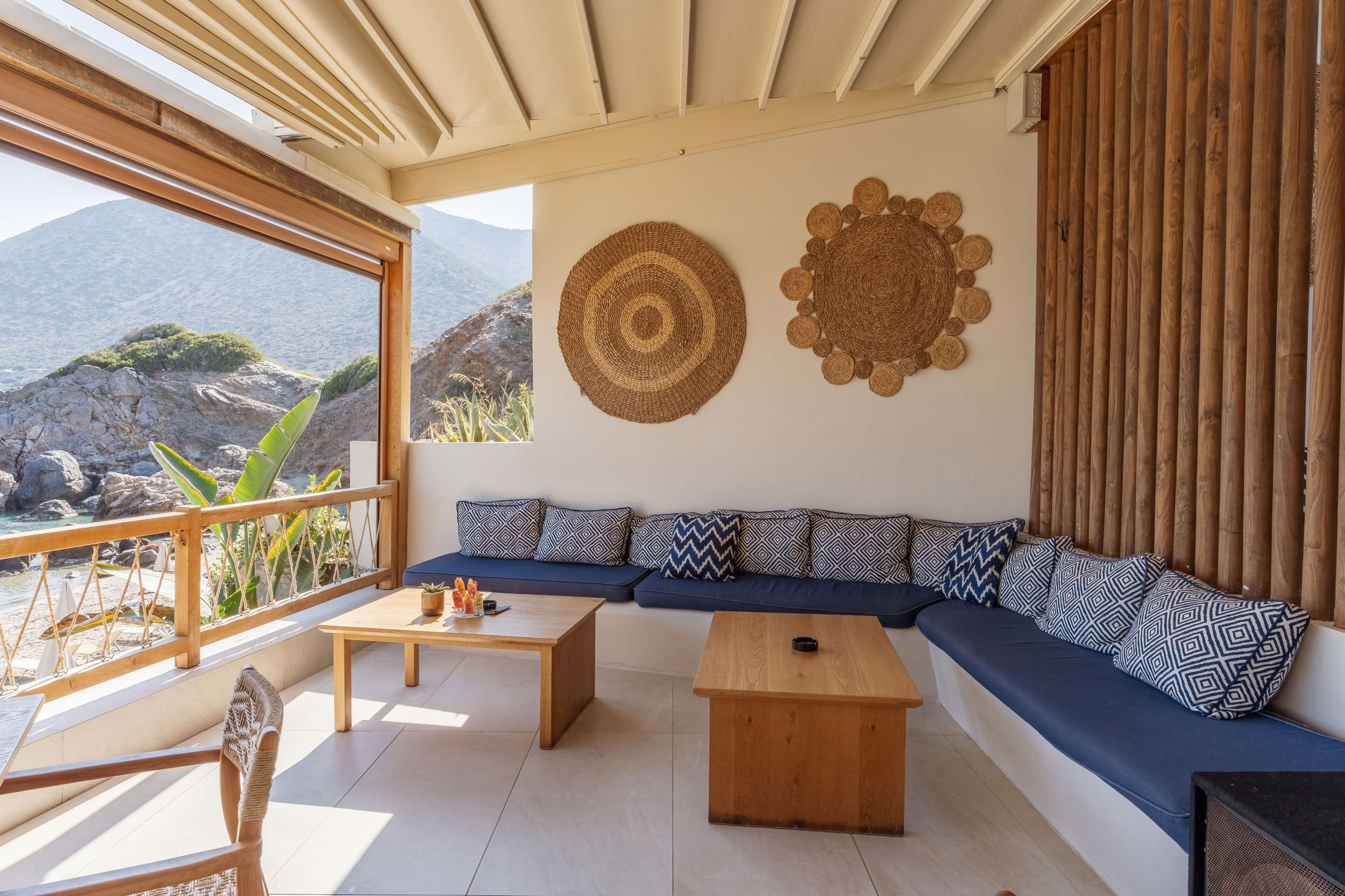 A modern living room with a large window overlooking a mountainous landscape, a wooden sofa with patterned cushions, a wooden coffee table, and a woven wall hanging.