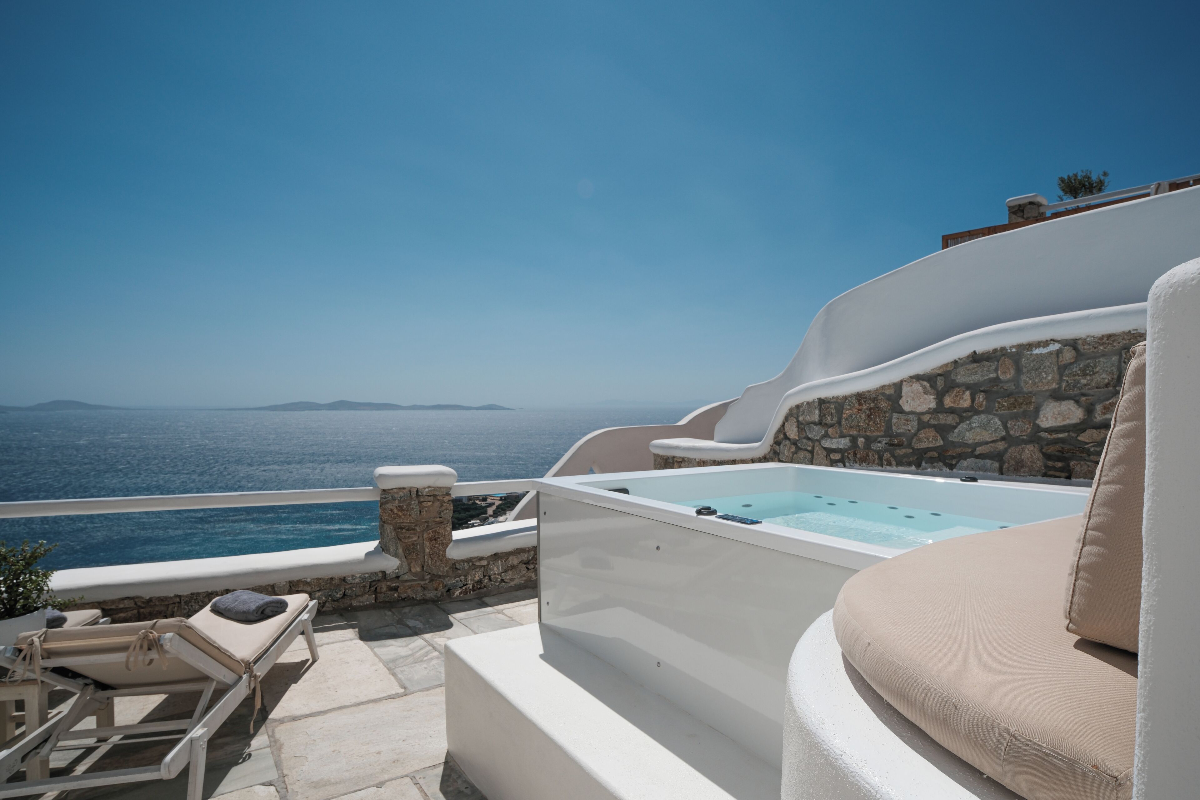 A sunlit outdoor area with a stone wall, a hot tub, lounge chairs, and a view of the sea.