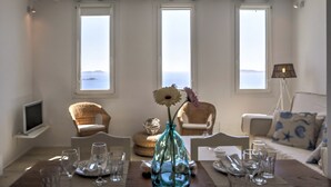 A dining area with a table set for four, three windows with a view of the sea, wicker chairs, and a lamp. - La Maison Blanche (Mykonos)