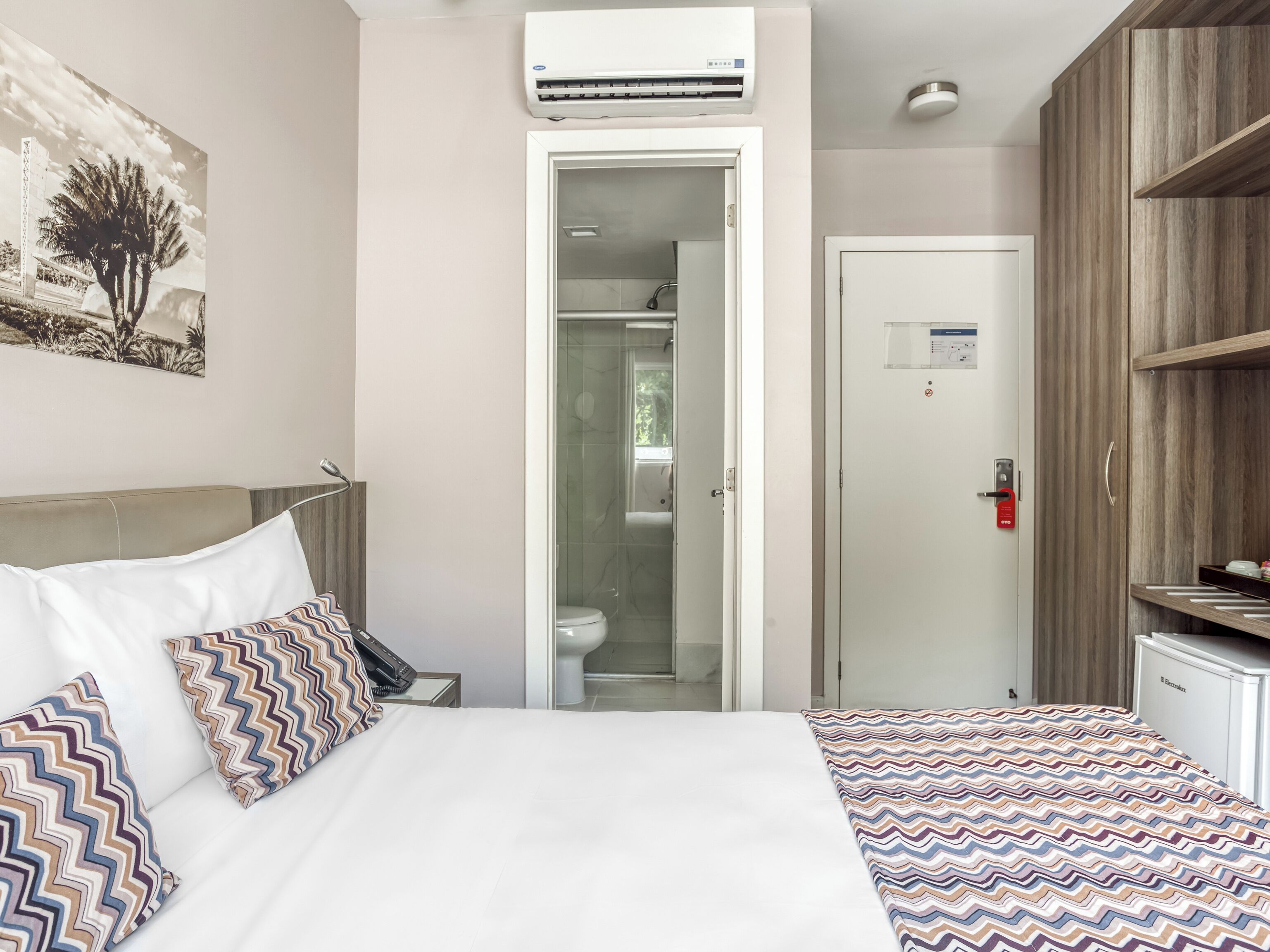 A hotel room with a bed, a nightstand, a telephone, a door to a bathroom, and a shelf with items.