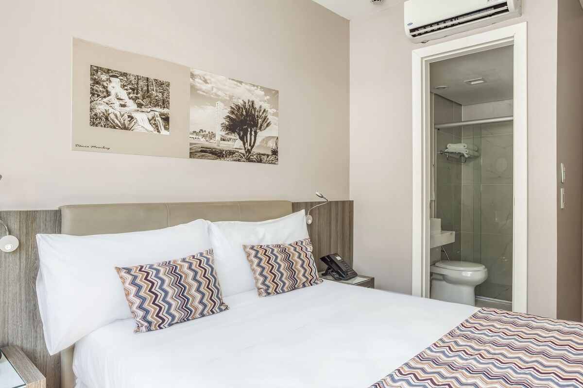 A hotel room with a bed, a bedside table with a lamp, a phone, and a wall-mounted air conditioner.