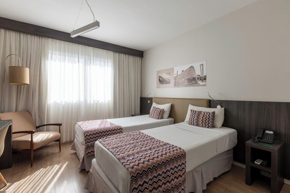 Ein Hotelzimmer mit zwei Betten, einem Schreibtisch, einem Stuhl, einer Lampe und einem Telefon auf einem Nachttisch.