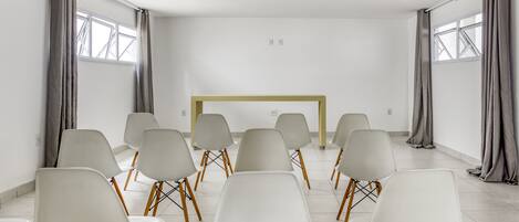 Un salón de conferencias con sillas blancas dispuestas en filas, una mesa amarilla y ventanas grandes con cortinas grises.