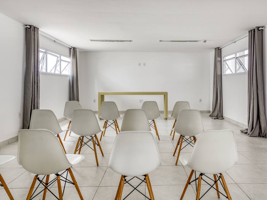 A conference room with white chairs arranged in rows, a yellow table, and large windows with grey curtains.