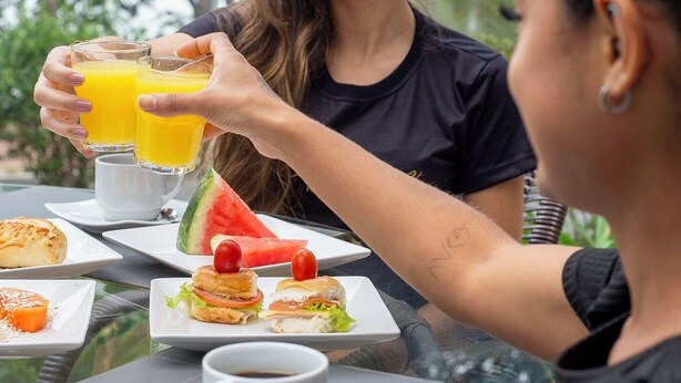 Una mesa de comedor con comida y bebidas, incluyendo un vaso de zumo de naranja, un plato de sándwiches y una taza de café.