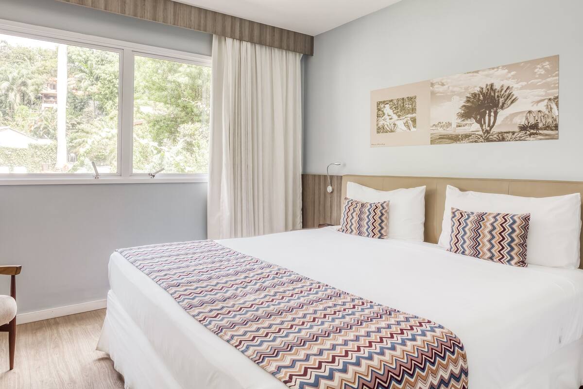 Ein Hotelzimmer mit einem großen Bett, einem Fenster mit Vorhängen, einem Wandbild und einem Holz-Kopfteil.