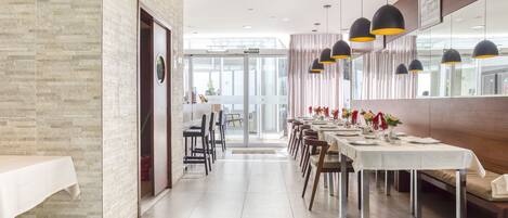 Un comedor moderno con una larga mesa dispuesta para una comida, una pared de ladrillo en un lateral y un vidrio que da a otra habitación.