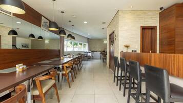 A modern restaurant with long wooden tables and black chairs, a wooden reception desk, and pendant lighting.