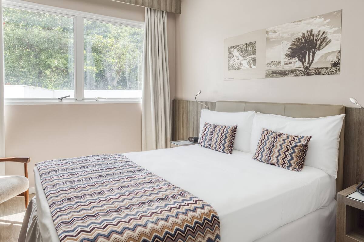 Ein Hotelzimmer mit einem großen Bett, einem Fenster mit Vorhängen, einem Nachttisch und einer Wandlampe.