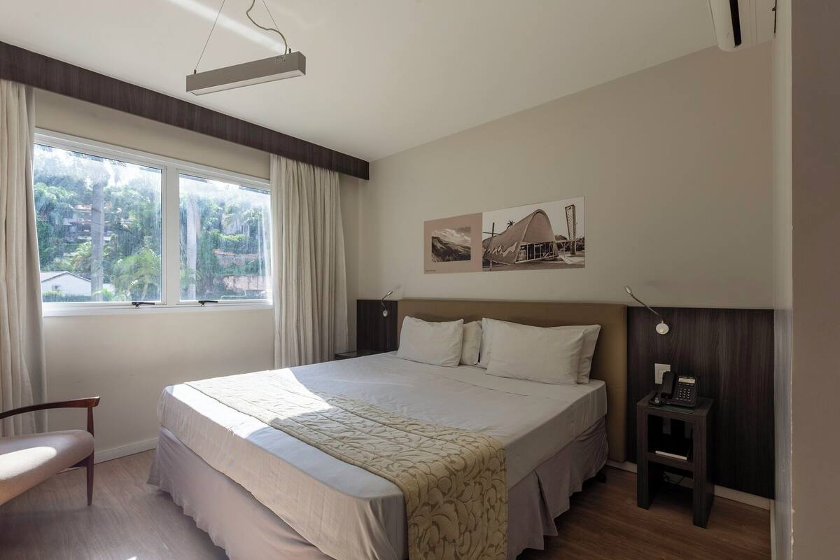 Ein Hotelzimmer mit einem großen Bett, einem Nachttisch mit Telefon, einem Sessel und einem Fenster mit Vorhängen.