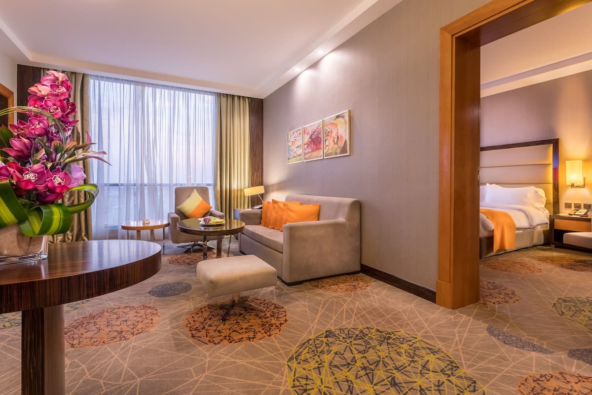 A hotel room with a sofa, armchair, and ottoman, a dining table, and a large window with curtains.
