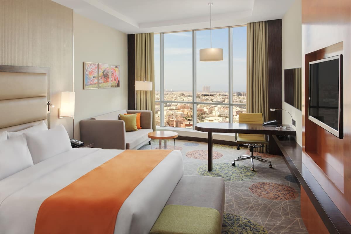 A hotel room with a large bed, a sofa, a desk, and a TV. There is a window with a view of the city.