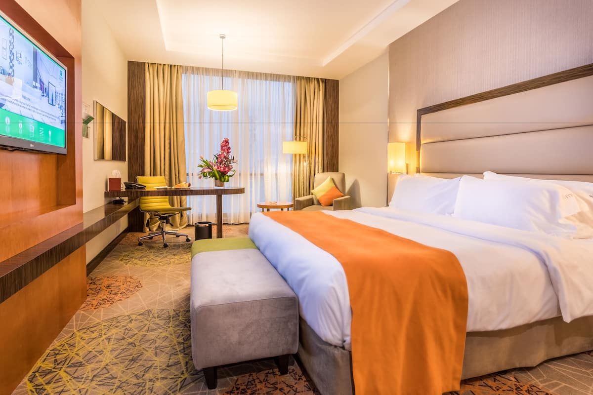 A hotel room with a large bed, a desk with a computer, a chair, and a small table with a vase of flowers.