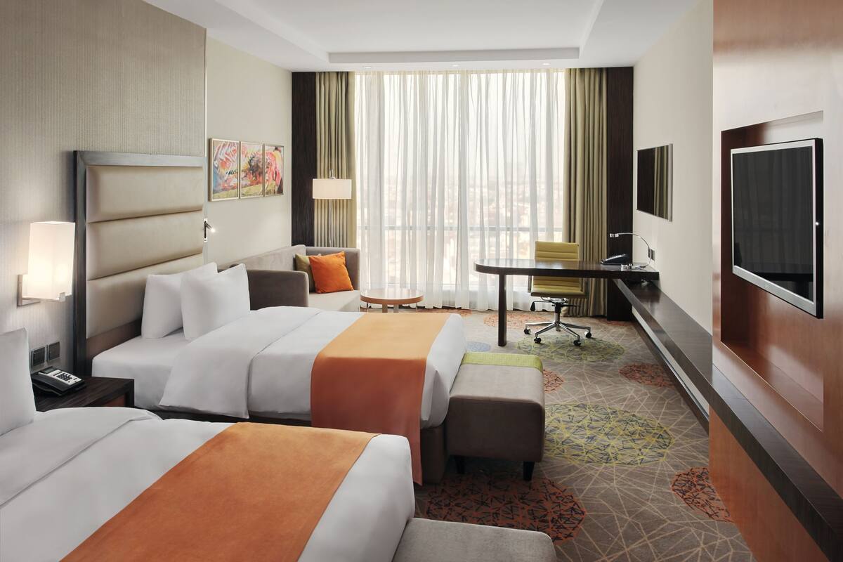 A hotel room with two beds, a desk, a chair, a TV, and a window with curtains.