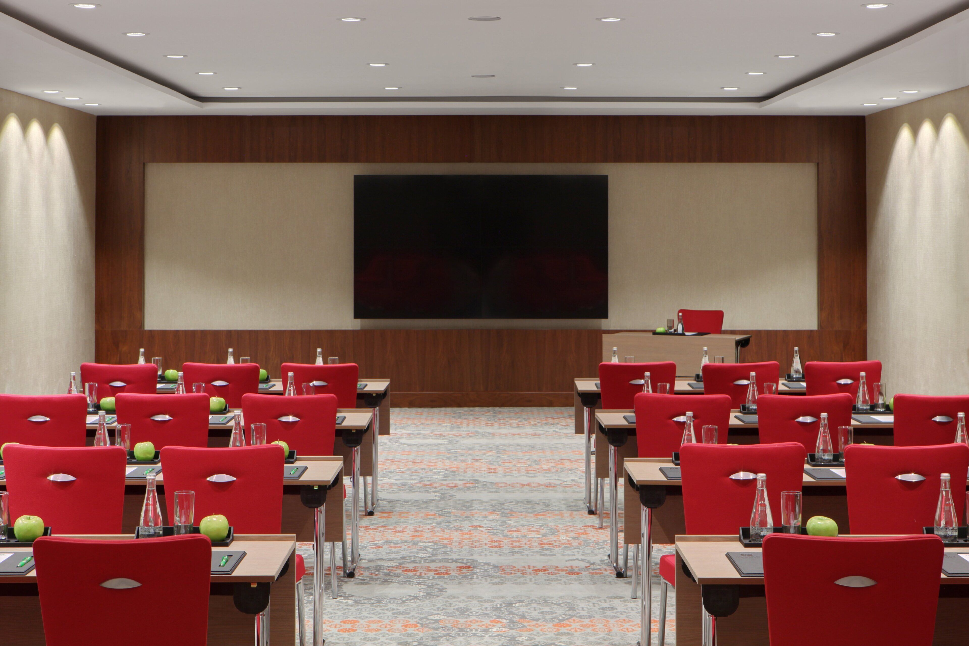 A conference room with rows of red chairs, tables with water bottles and apples, and a large screen on the wall.