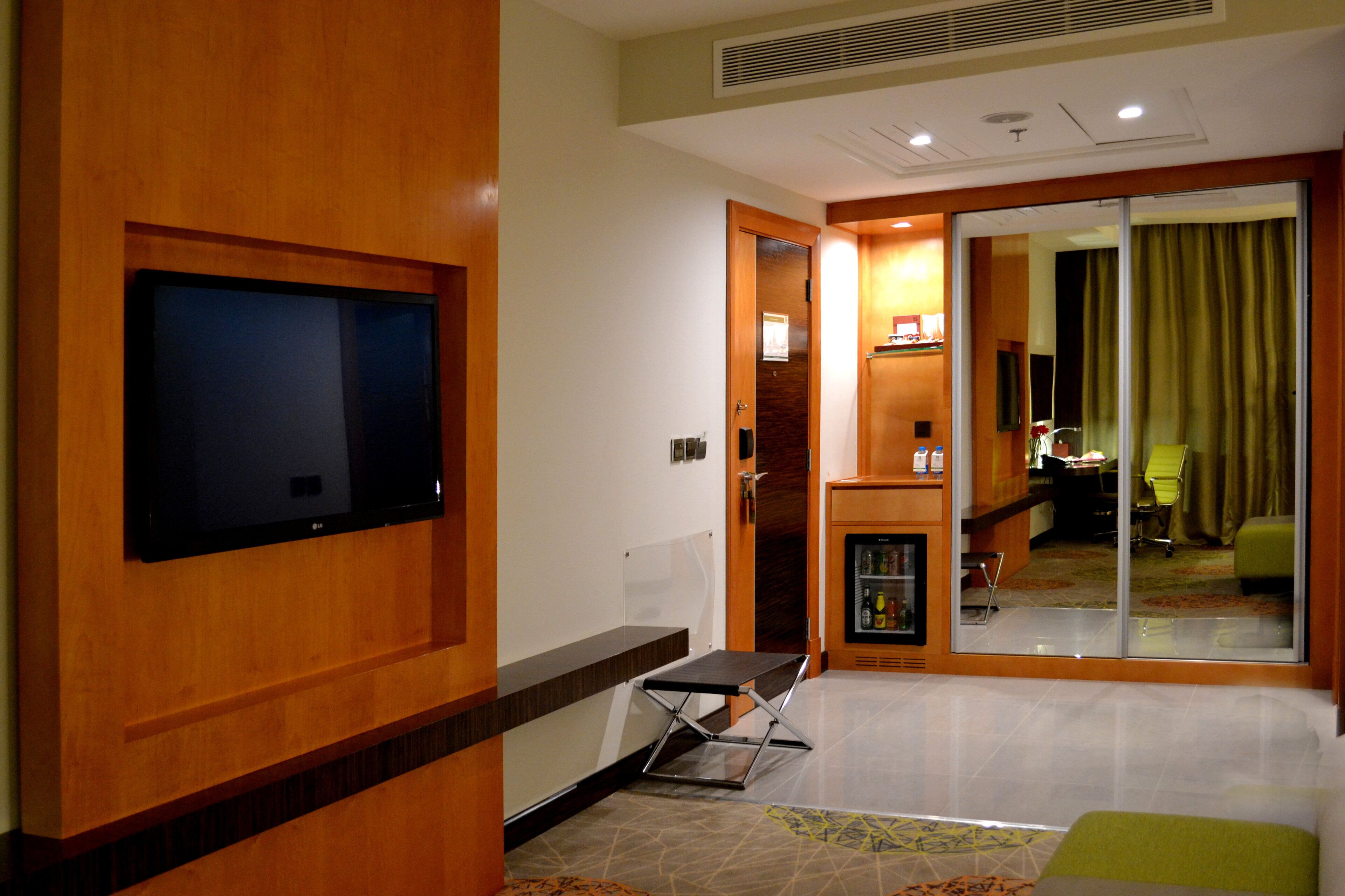 Habitación de hotel con una televisión de pantalla plana montada en la pared, una mesita con una silla, un refrigerador y un ventanal corredero que da a un balcón.