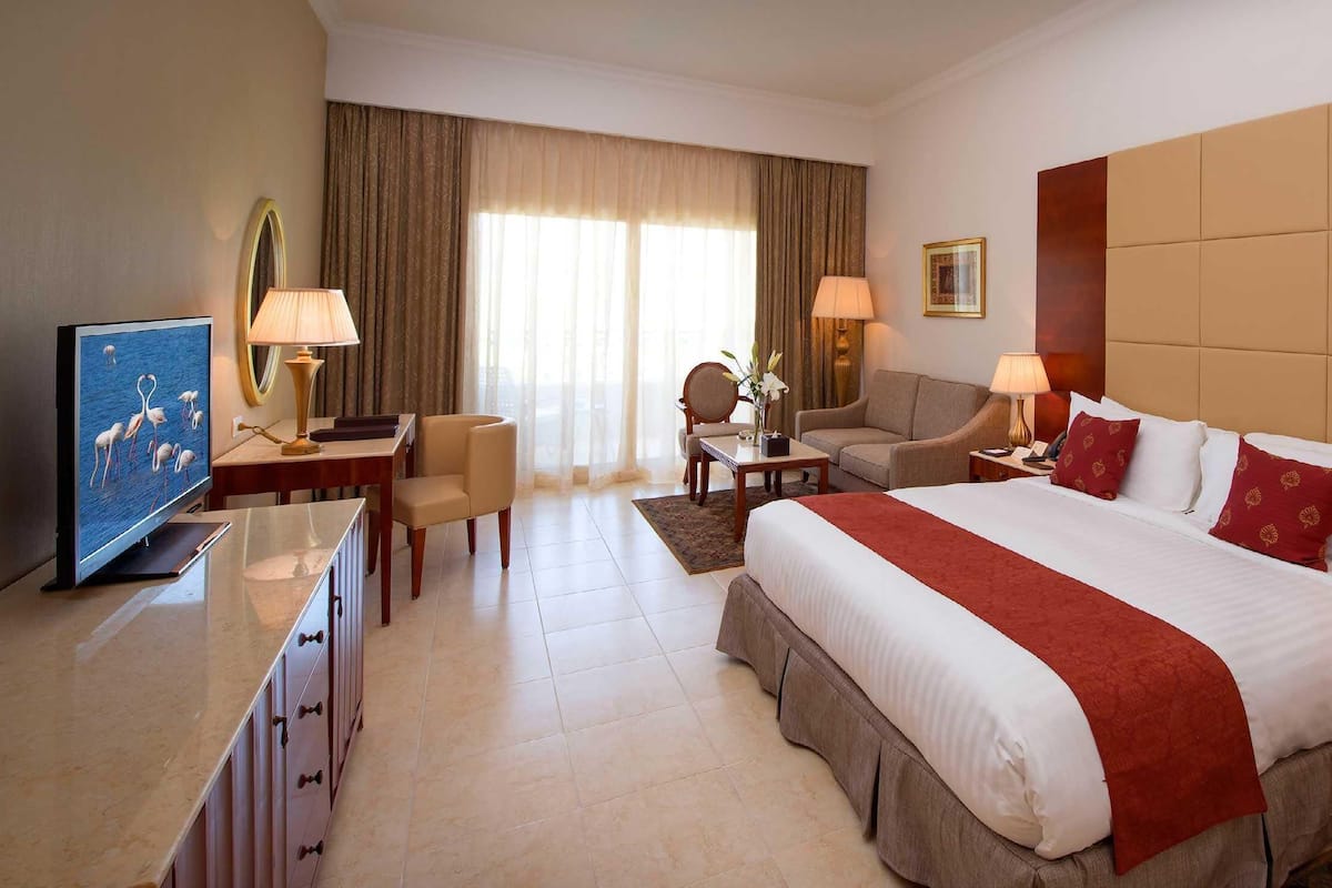 A hotel room with a large bed, two bedside tables, a desk with a chair, a television, and a view of the outdoors through the window.