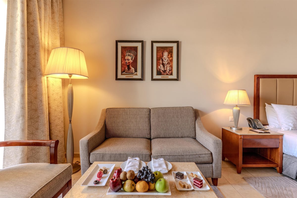 A hotel room with a sofa, a dining table with fruit and pastries, a lamp, and two framed pictures on the wall.