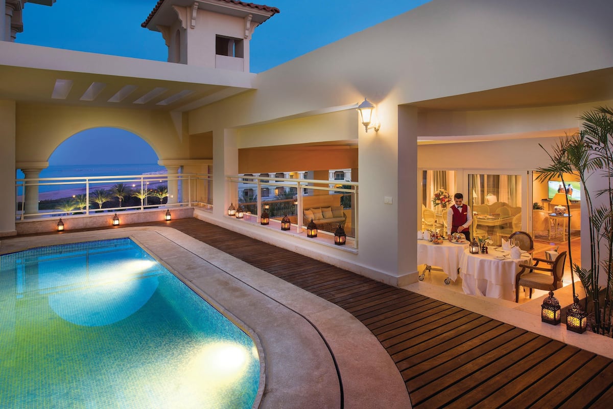A pool area with a view of the ocean, outdoor dining with tables and chairs, and a service area with a staff member.