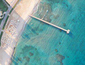 An aerial view of a beach with a wooden pier extending into the turquoise water, surrounded by clear shallow areas and small islands.