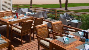 An outdoor seating area with wooden tables and chairs, overlooking a beach with palm trees and the ocean. - Baron Palace Sahl Hasheesh (Sahl Hasheeh)