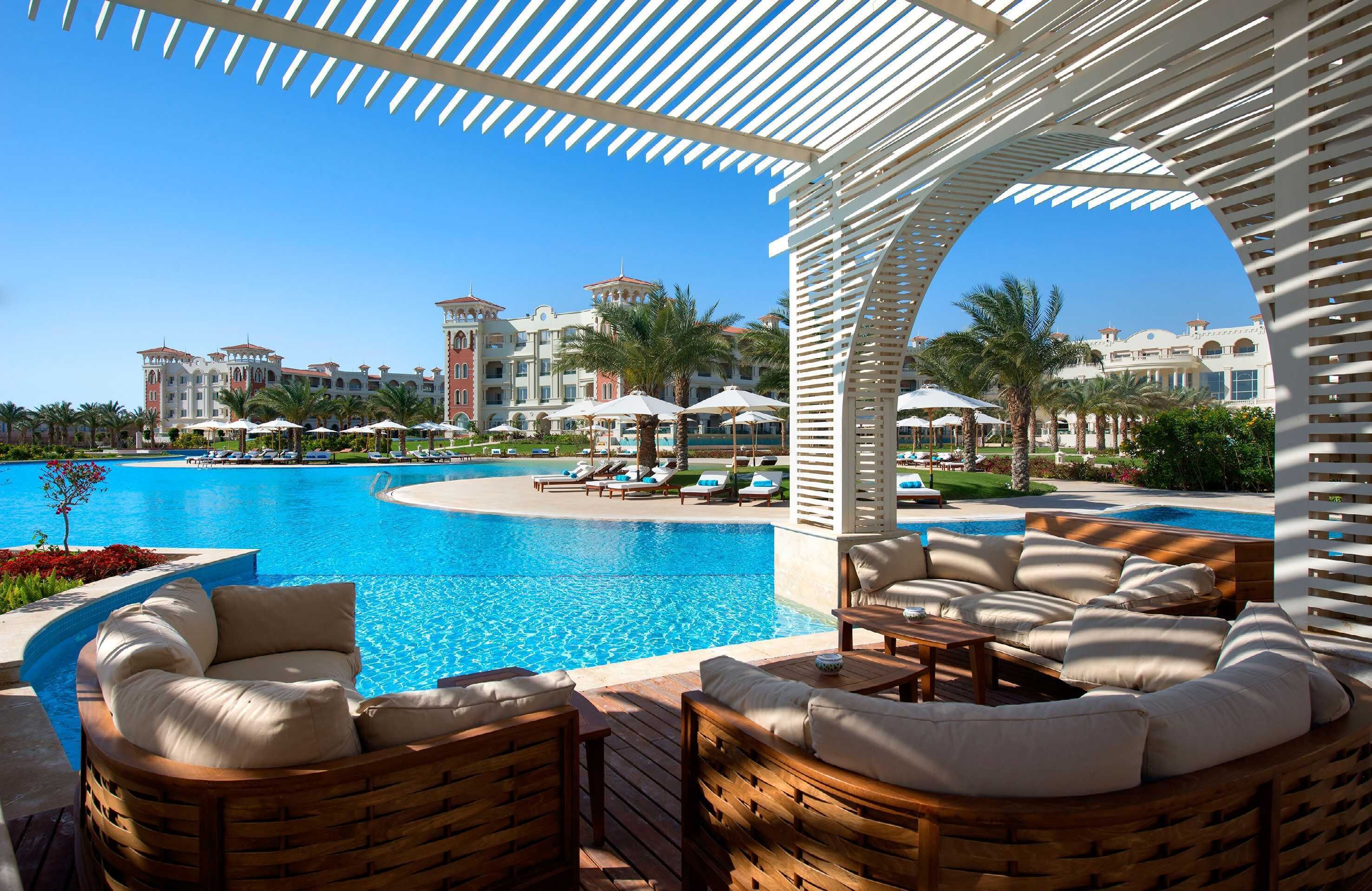 Un espace aménagé au bord de la piscine, avec des chaises longues et des parasols, une piscine à l’eau d’un bleu limpide et, au fond, le bâtiment de l’hôtel.