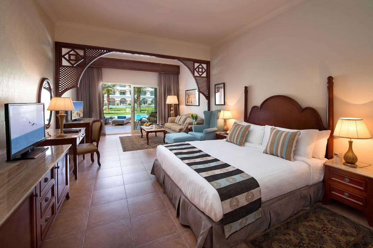 A hotel room with a large bed, a desk with a TV, a chair, a sofa, and a view of the outdoor pool area.