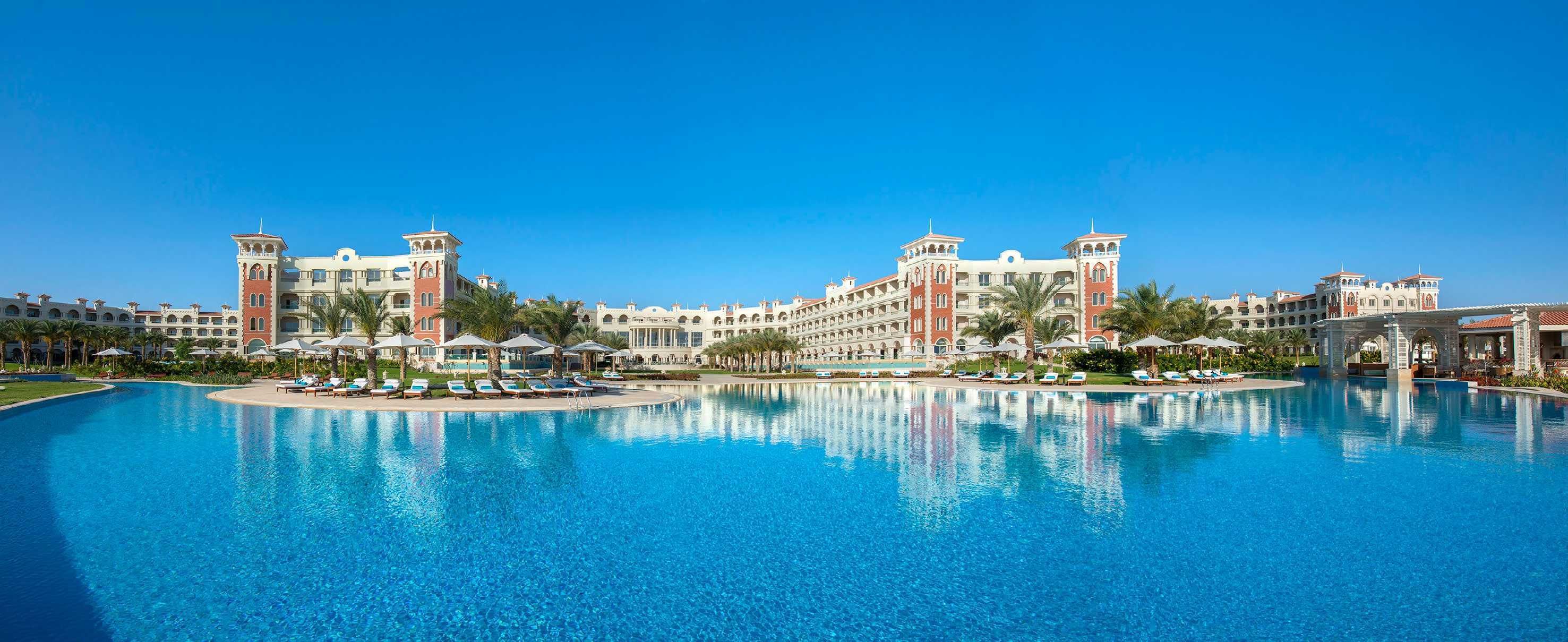 Una piscina grande con sillones de descanso y sombrillas, rodeada por un hotel con varias edificaciones y palmeras.