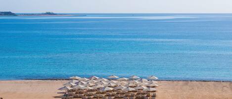 A beach with rows of sun loungers and umbrellas, a clear blue sea, and a distant shoreline with buildings.