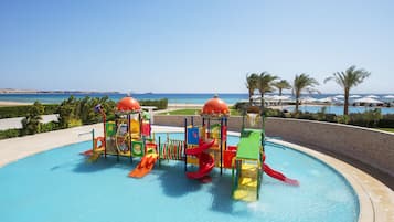 Children can play and swim in a vibrant pool area surrounded by a beach and palm trees.