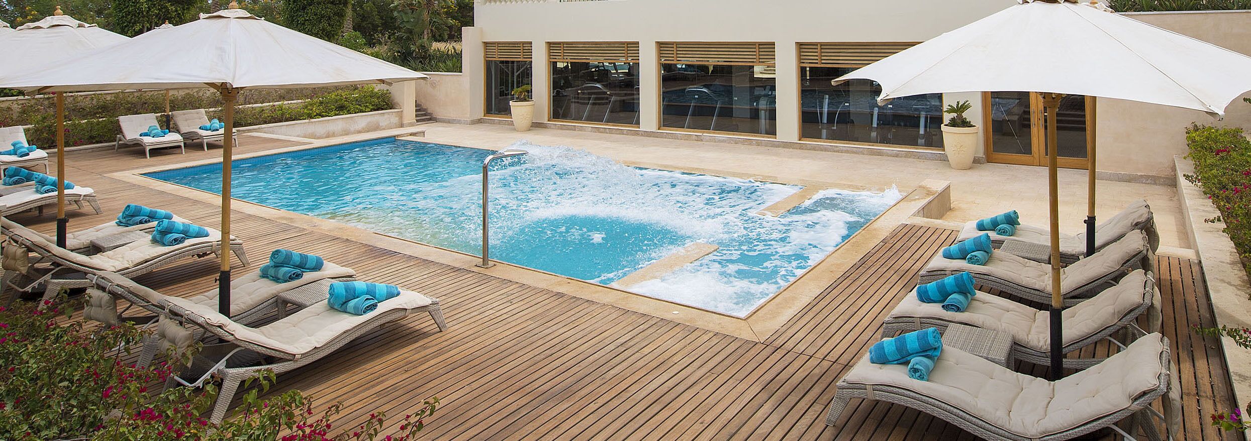 Un área de piscina con sillones de descanso y sombrillas, un jacuzzi y un edificio con grandes ventanales.