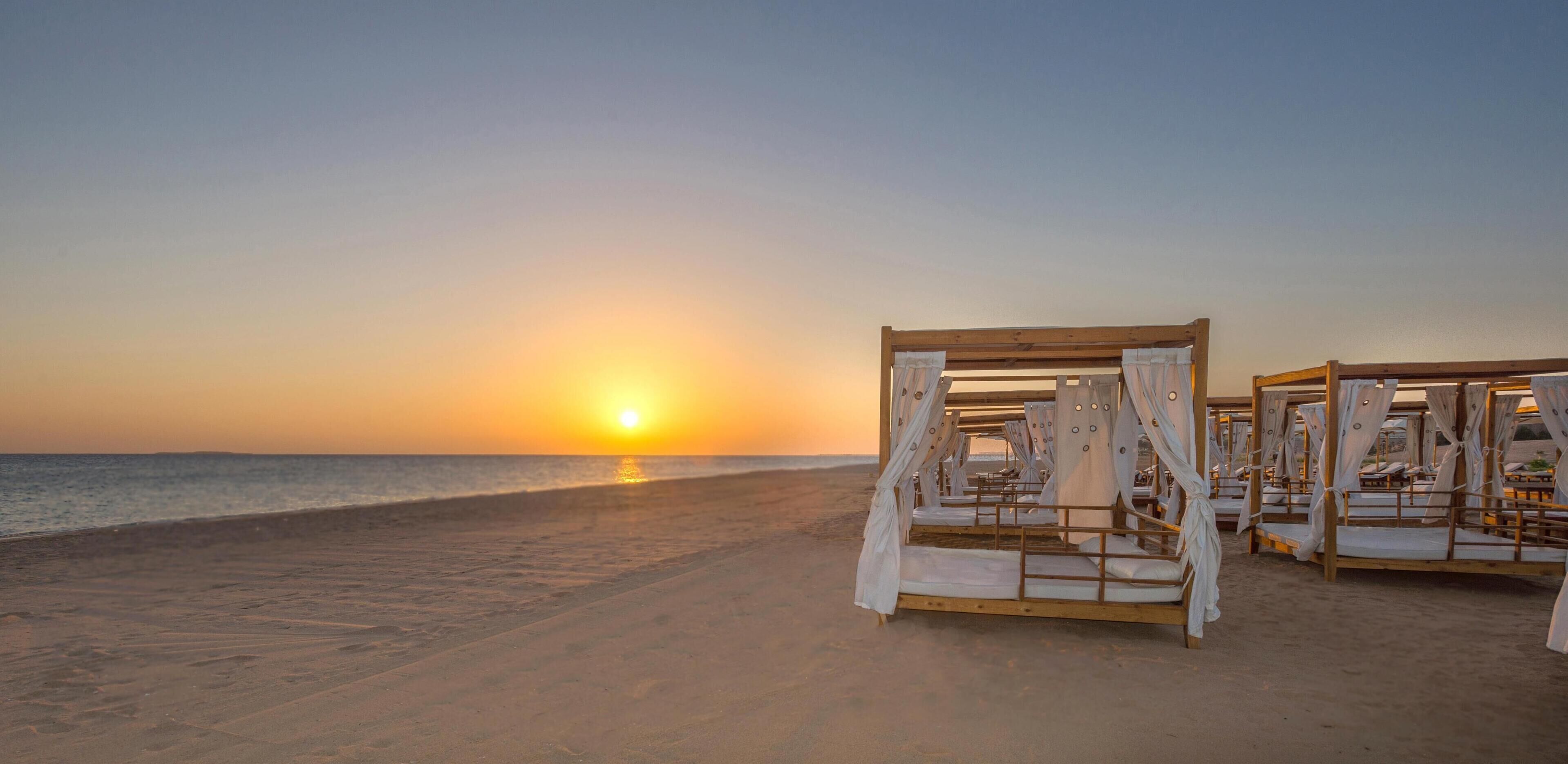 Una playa con camas de madera y cortinas blancas, con un atardecer de fondo.