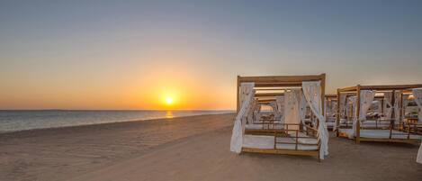 Una playa con camas de madera y cortinas blancas, con un atardecer de fondo.