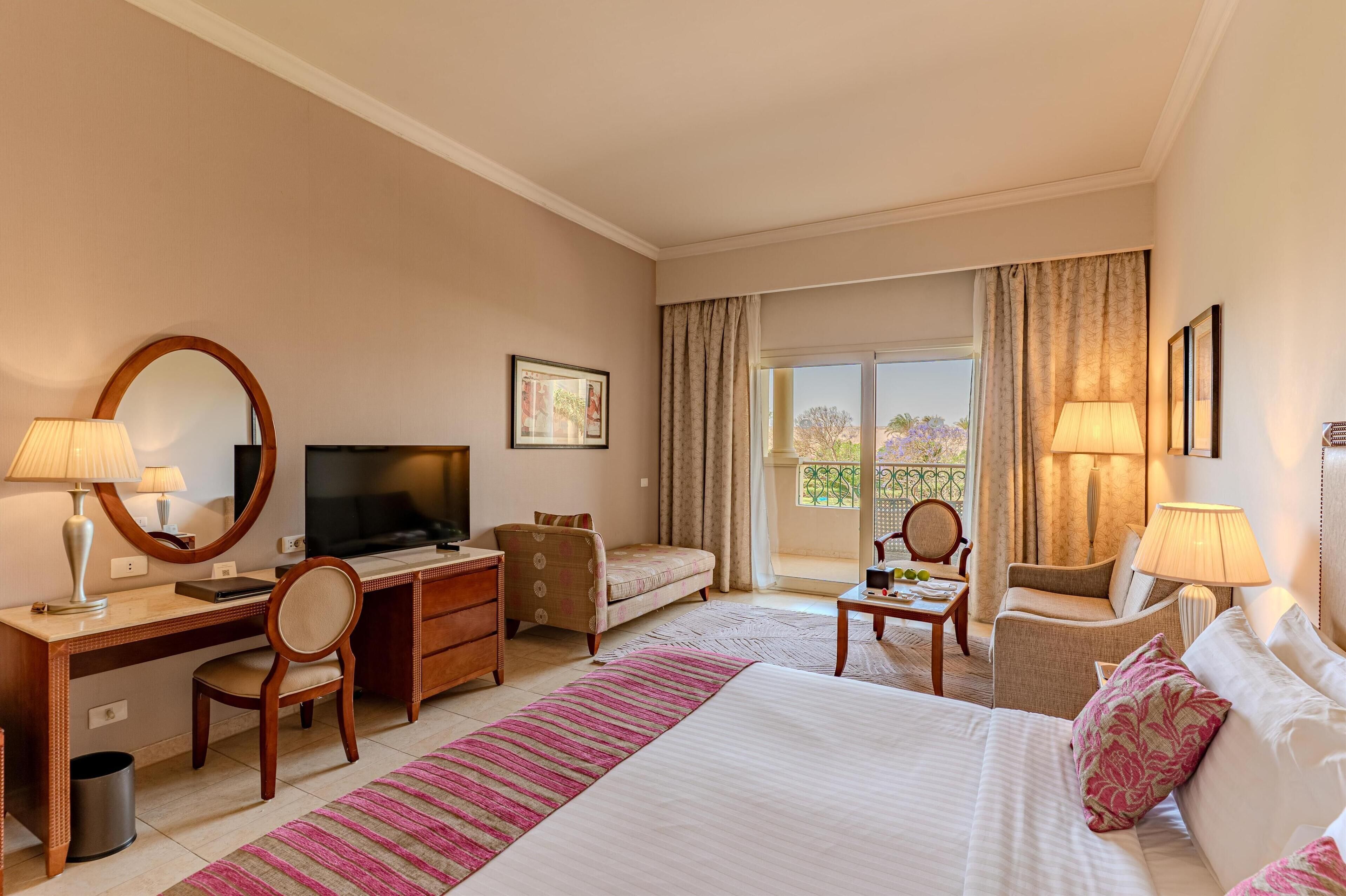 Habitación de hotel con cama, escritorio, silla, televisor y balcón con cortinas.