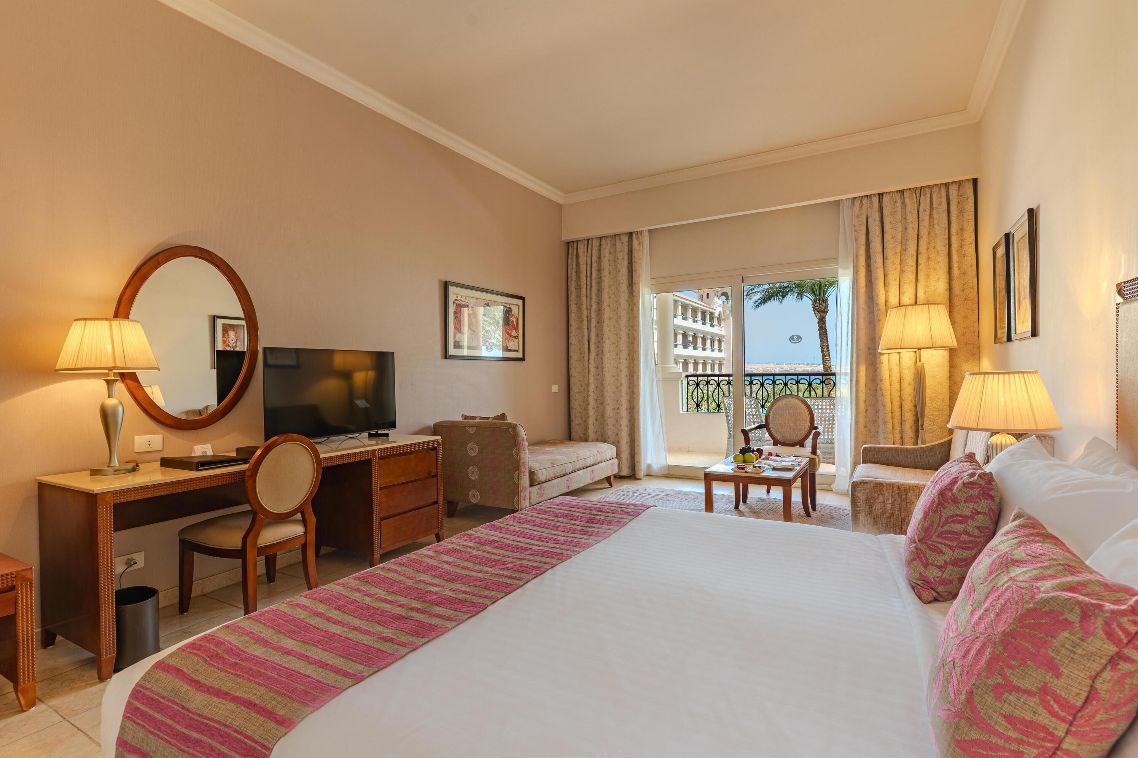 Habitación de hotel con cama, escritorio, silla, televisor y balcón con cortinas.