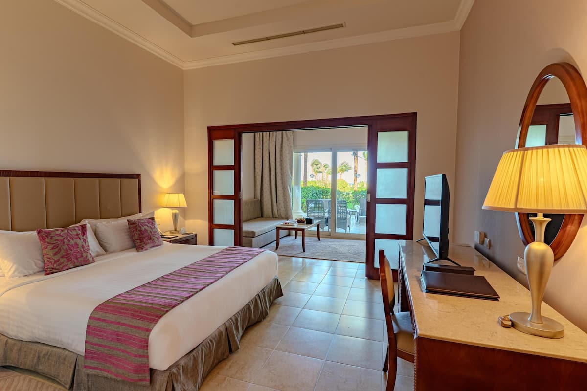 A hotel room with a large bed, a desk, a chair, a sofa, and a balcony with a view of palm trees.